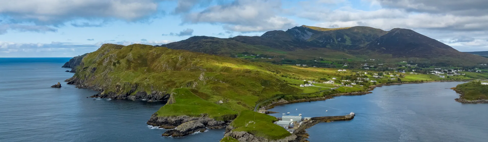 Teelin Harbour's boat dock, and high, cloud-covered mountains in the background under a partly cloudy sky.